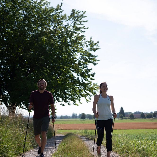 Ein Paar betreibt Nordic Walking auf einem Feldweg in ländlicher Landschaft.