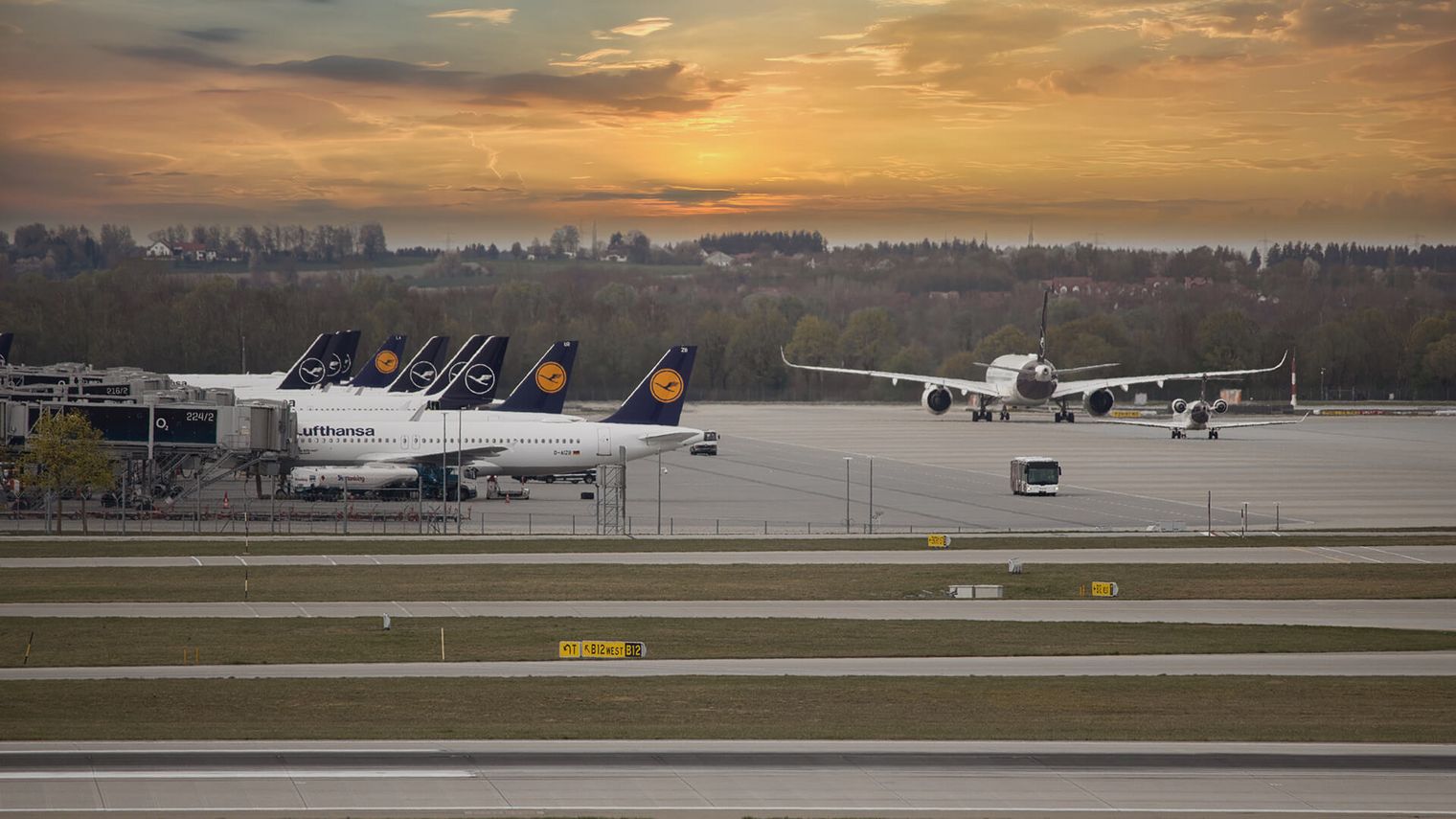 Mehrere Flugzeuge auf dem Rollfeld des Flughafens München bei Sonnenuntergang, mit geparkten Maschinen und einem startbereiten Flugzeug im Hintergrund.