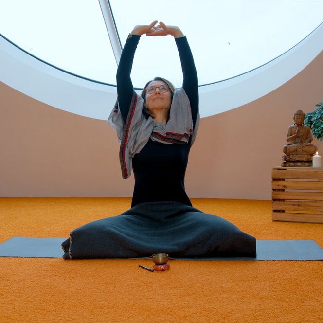 Eine Frau sitzt auf einer Yogamatte in einem Ruheraum und streckt die Arme nach oben; daneben stehen Buddhafigur, Kerzen und eine Pflanze.