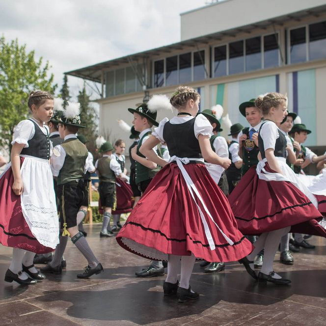 Kinder tanzen in rot-weißen Trachten auf einer Bühne.
