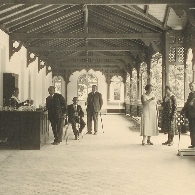 Historische Schwarz-Weiß-Aufnahme einer Wandelhalle im Kurpark mit Gästen, Sitzbänken, Ausschank und verzierten Holzarkaden.