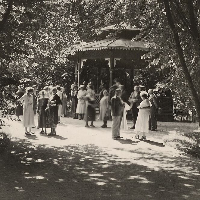 Historische Schwarz-Weiß-Aufnahme: Menschen versammeln sich vor einem Pavillon im schattigen Kurpark.