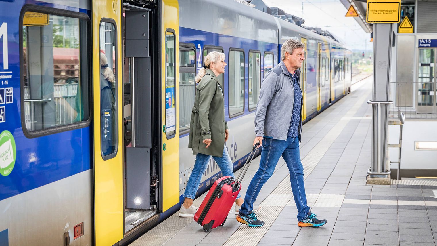 Ein Paar steigt mit Gepäck aus einem Zug auf einem Bahnsteig aus, bereit zur Weiterreise.