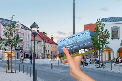Hand hält die Bad Aiblinger Gästekarte hoch, im Hintergrund sieht man den belebten Marienplatz im Stadtzentrum bei sommerlicher Abendstimmung