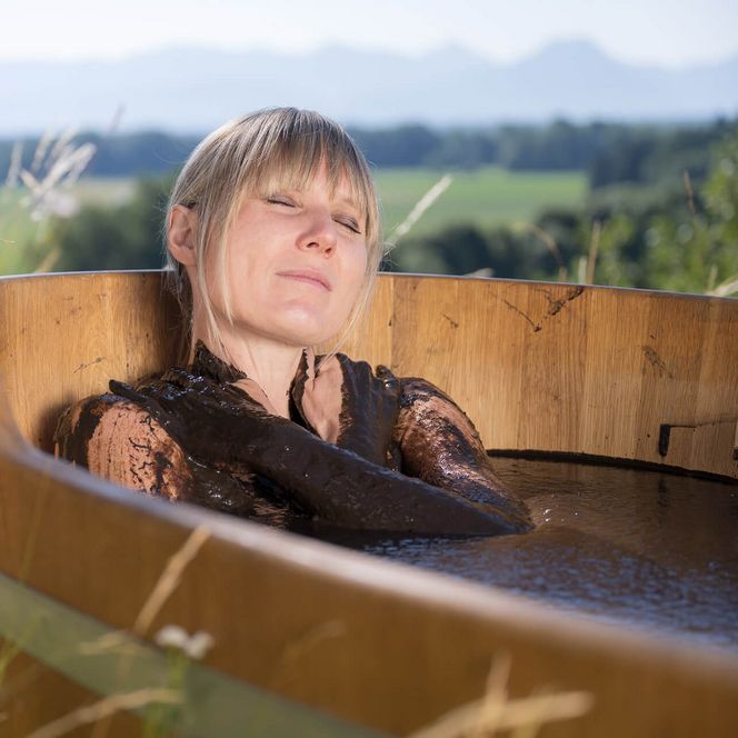 Frau entspannt mit geschlossenen Augen in einer Moorwanne auf einer Wiese vor weiter Landschaft.