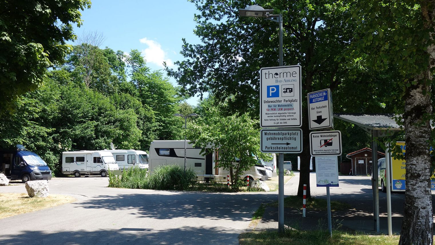Wohnmobilstellplatz an der Therme Bad Aibling mit mehreren geparkten Wohnmobilen, umgeben von Bäumen und Hinweisschildern zum gebührenpflichtigen Parken.