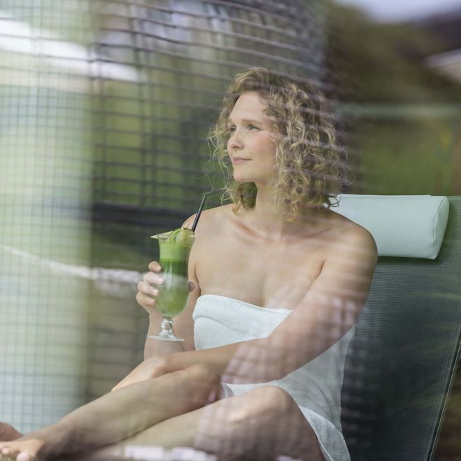 Eine Frau entspannt in einer Therme auf einer Liege und hält ein grünes Getränk in der Hand.