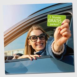 Eine lächelnde Person sitzt in einem Auto und hält eine grüne Karte mit dem Text „1 Stunde GRATIS parken“ aus dem Fenster.