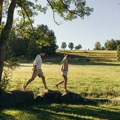 Ein Paar balanciert barfuß über Steine auf einer sonnigen Wiese in ländlicher Natur.