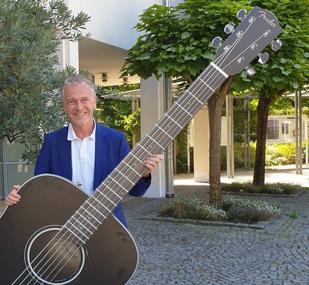 Ein lächelnder Mann im blauen Sakko hält im Freien eine übergroße Fender-Gitarre vor Bäumen und einem Gebäude.