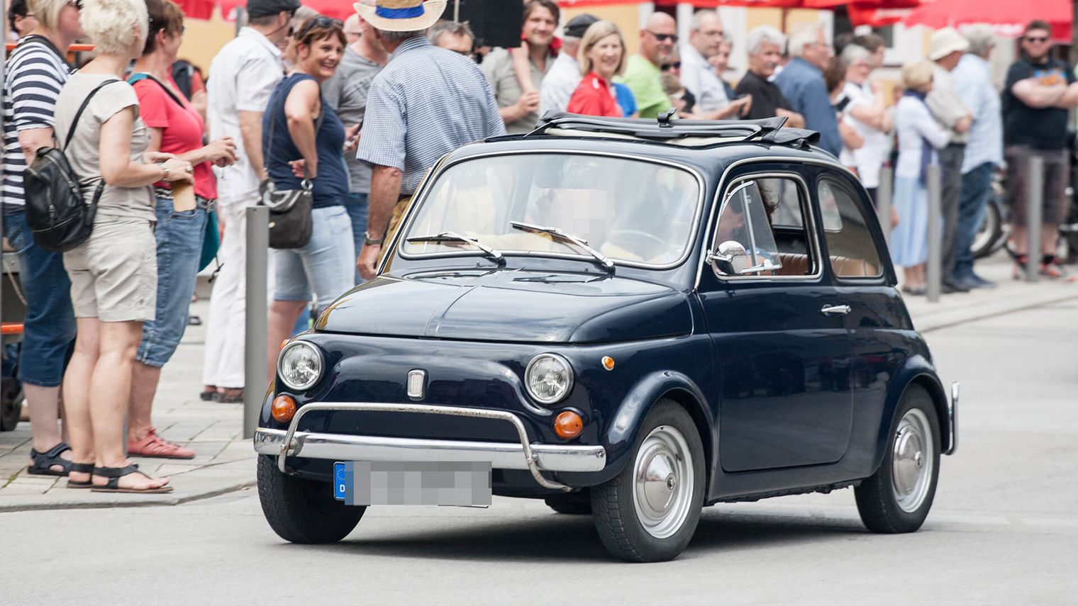 Kleines blaues Oldtimer-Auto fährt durch eine belebte Straße mit vielen Zuschauern am Rand.