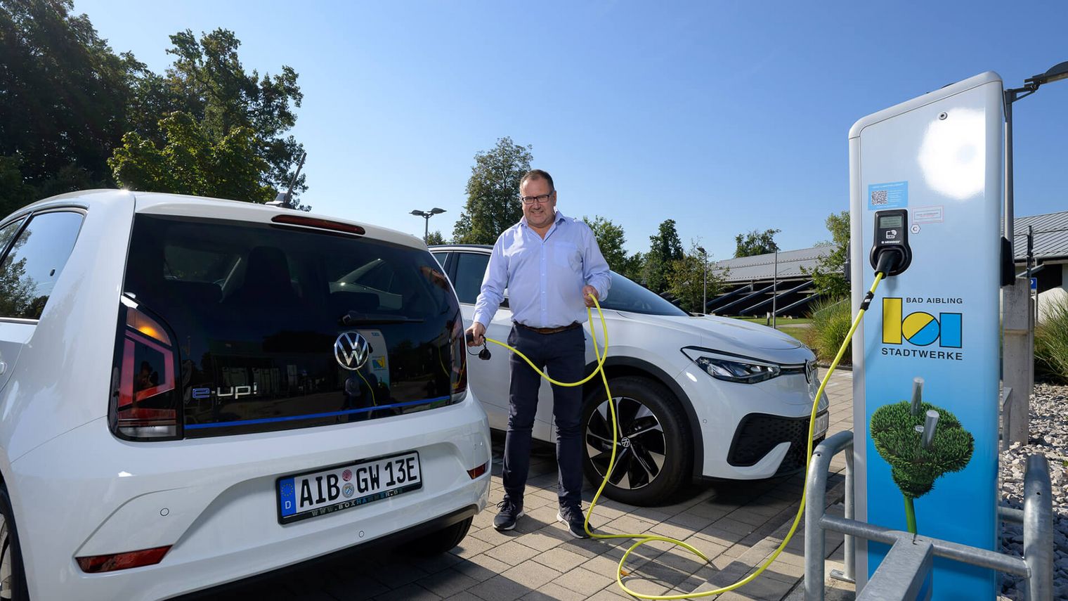 Ein Mann schließt ein Elektroauto an eine Ladesäule der Stadtwerke Bad Aibling an. Neben ihm stehen zwei weiße Fahrzeuge auf einem Parkplatz bei sonnigem Wetter.